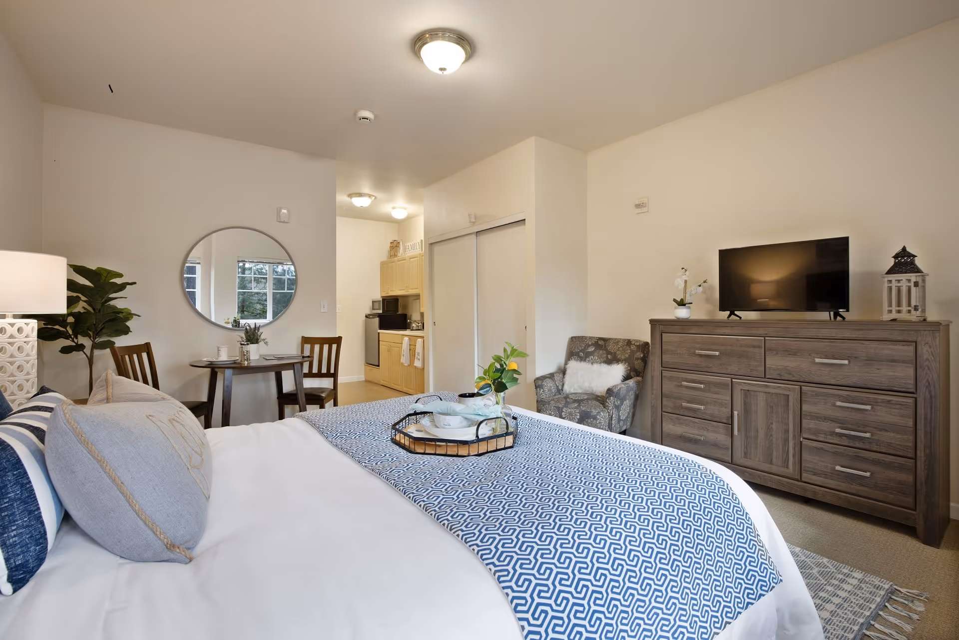 A cozy studio apartment featuring a neatly made bed with blue and white bedding, a small round dining table with two chairs, a patterned armchair, a wooden dresser with a flat-screen TV on top, and a kitchenette in the background. The room has neutral walls and a ceiling light fixture.