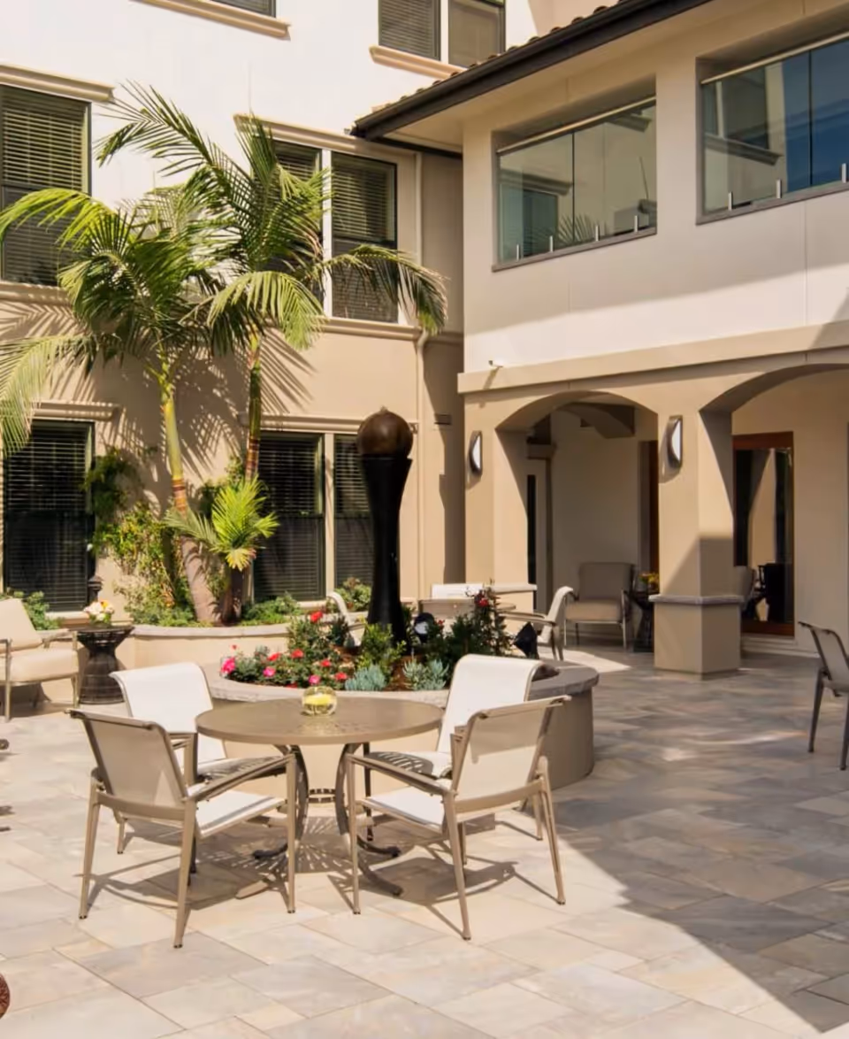 Sunlit courtyard with a round table and four chairs around a central planter/fountain, palm trees, and surrounding multi-story building windows.