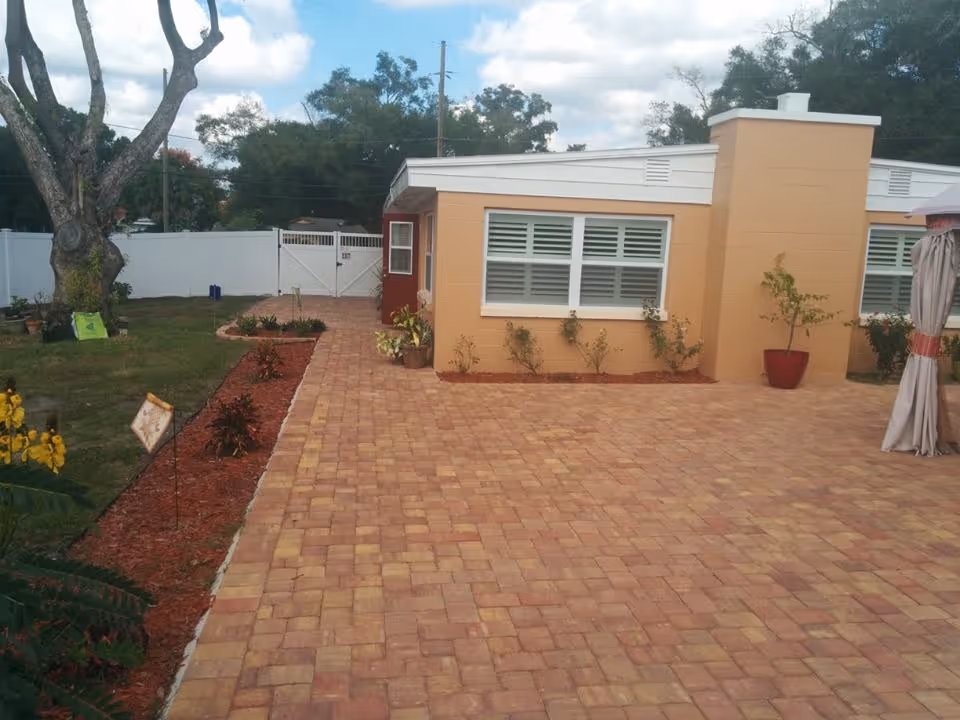 Outdoor view of a single-story building with beige and red walls, large windows with white shutters, and a paved brick patio. There are small plants and shrubs along the building and a large tree on the left side near a grassy area. A white fence and gate are visible in the background under a partly cloudy sky.