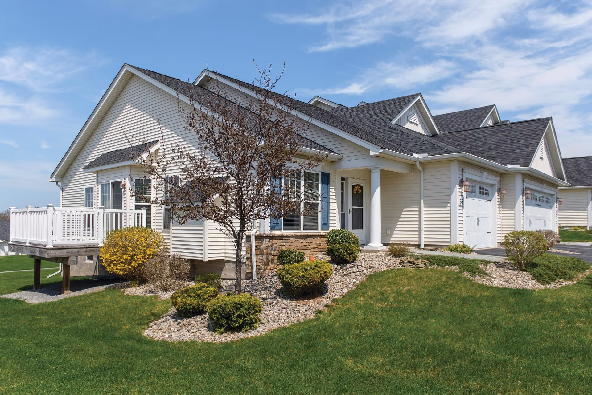 Exterior view of a single-story residential building with beige siding, a dark shingled roof, white garage doors, and a small porch supported by white columns. The front yard features green grass, small bushes, and a tree with sparse leaves under a partly cloudy blue sky.