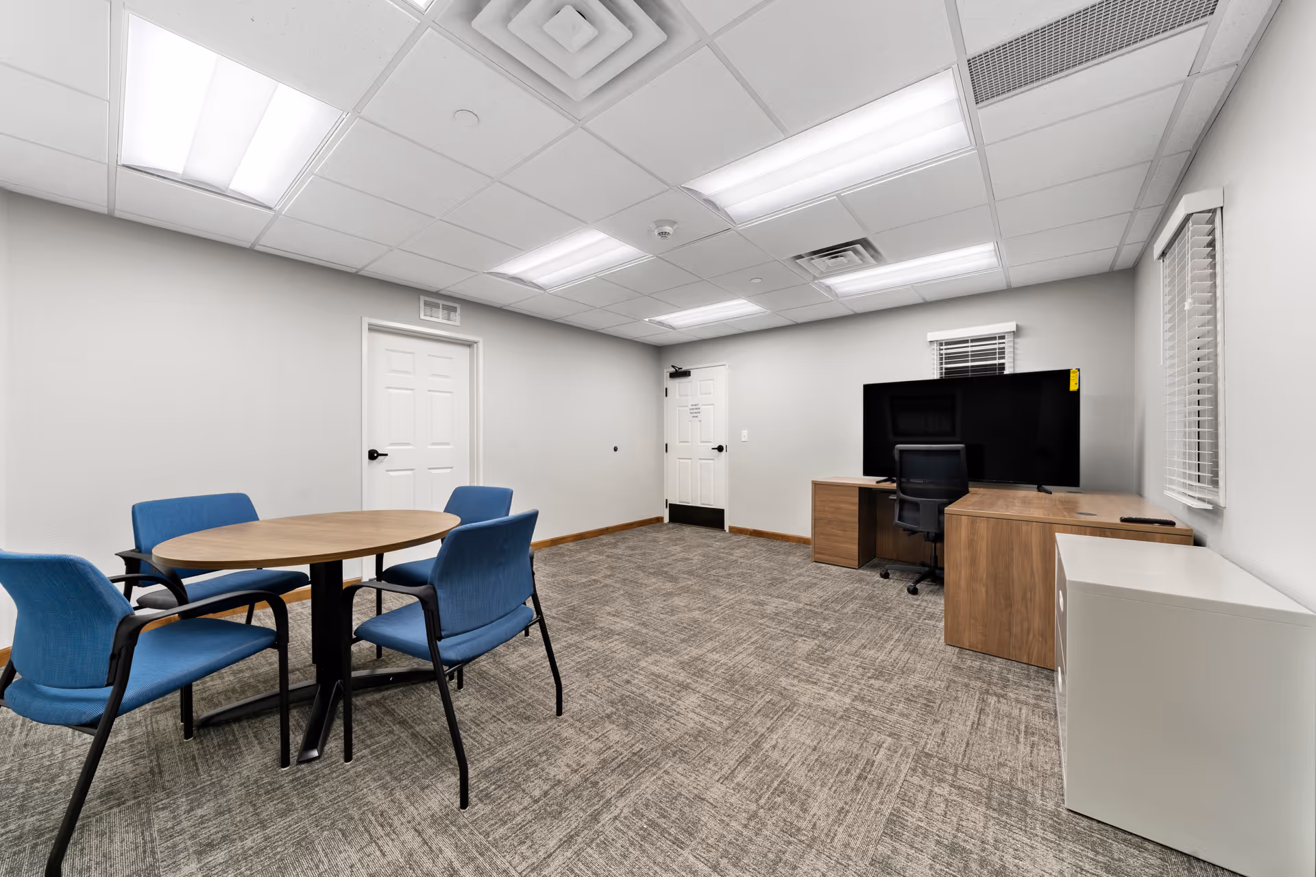 A small meeting or activity room with a round wooden table surrounded by four blue chairs on the left side. On the right side, there is a wooden desk with a black office chair and a large flat-screen TV on top. The room has gray carpet flooring, white walls, two white doors, and a ceiling with fluorescent lights and ventilation panels.