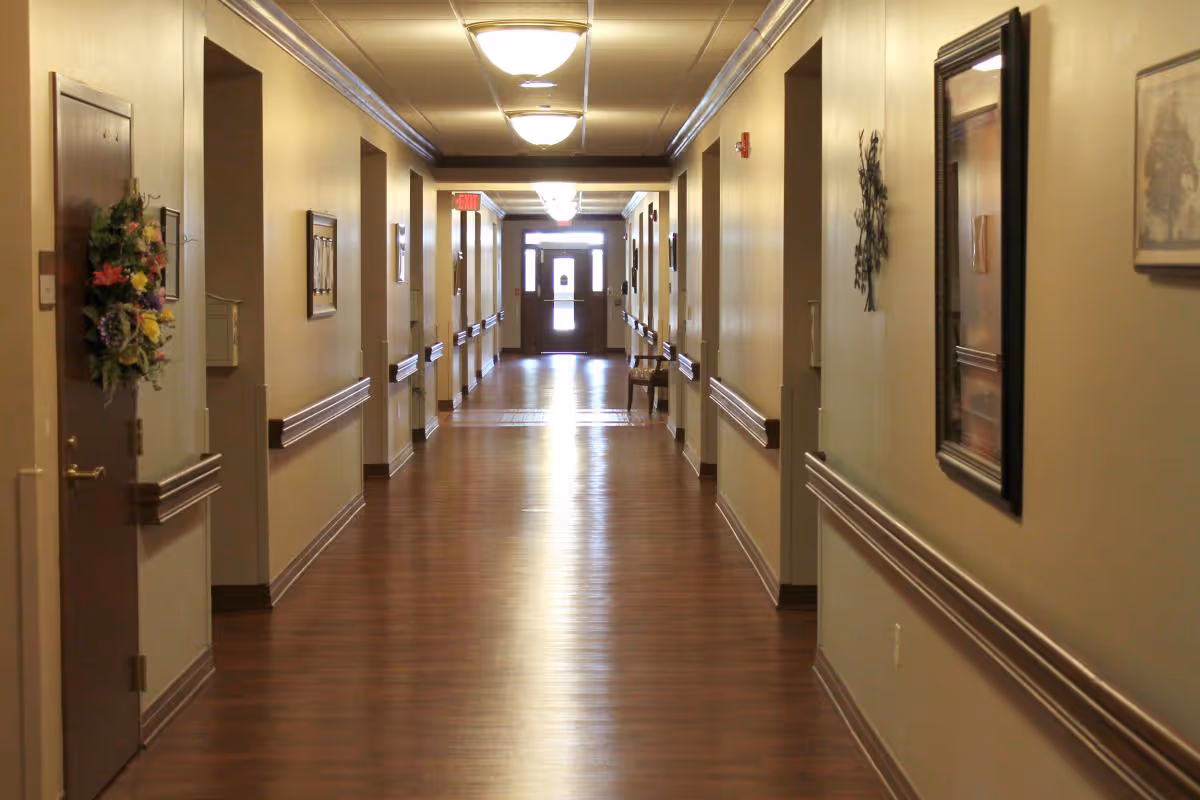 A long, well-lit hallway in a senior living facility with wooden floors and beige walls. Doors line both sides of the corridor, some decorated with wreaths and artwork. Handrails run along the walls, and ceiling lights illuminate the space. An exit door is visible at the far end of the hallway.