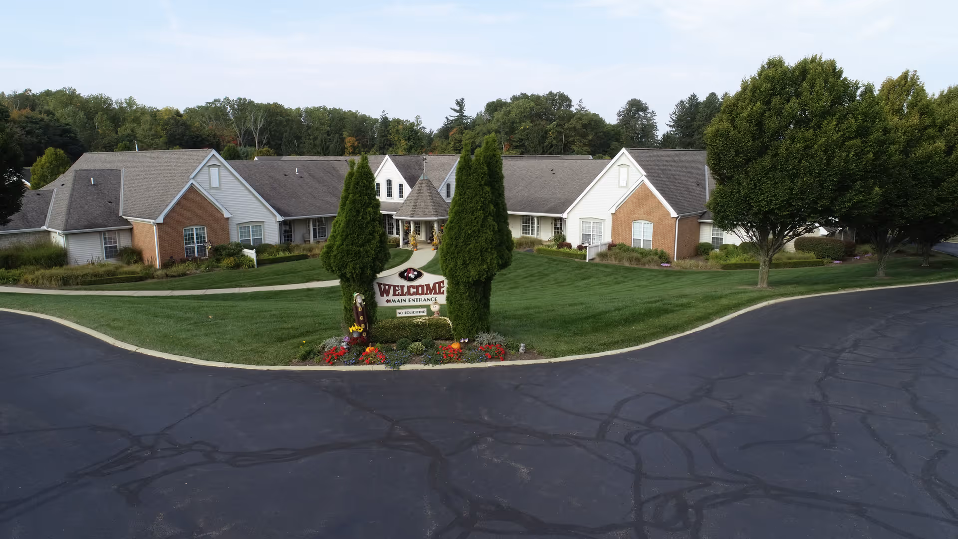 Exterior view of Wedgewood Estates senior living facility showing a single-story building with a mix of brick and siding, surrounded by well-maintained green lawns, trees, and a paved driveway. A welcome sign is placed in front of the building between two tall evergreen trees.