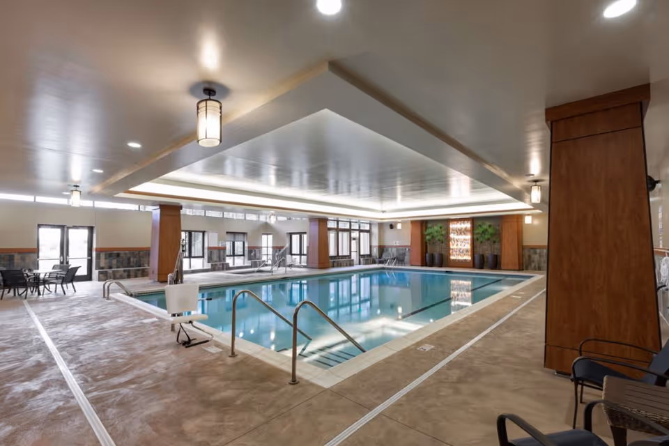 Indoor swimming pool area with seating, a pool lift, and large windows in a senior living facility.
