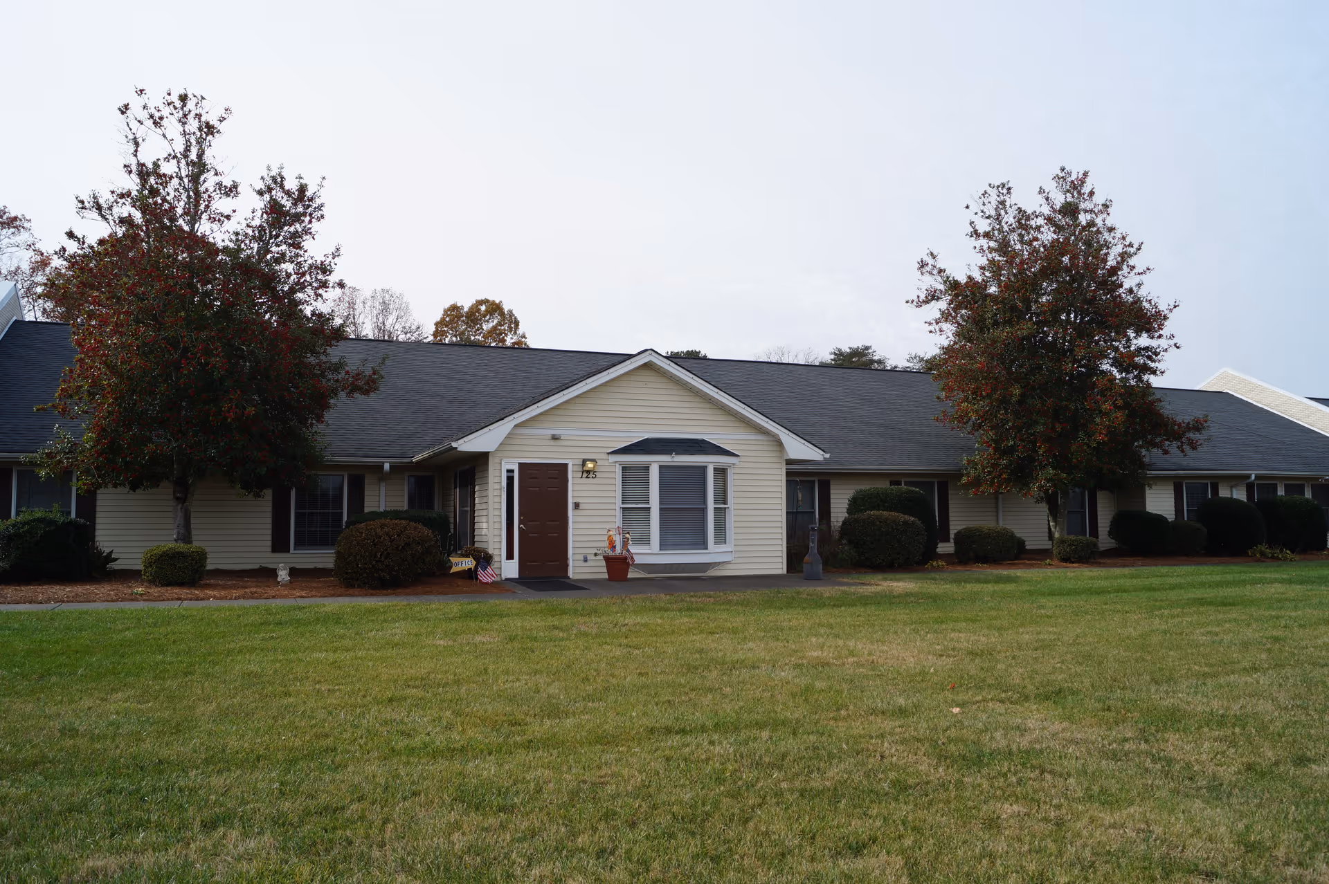 Front exterior of a single-story senior living building with a central entrance, bay window, shrubs, and a grassy lawn.