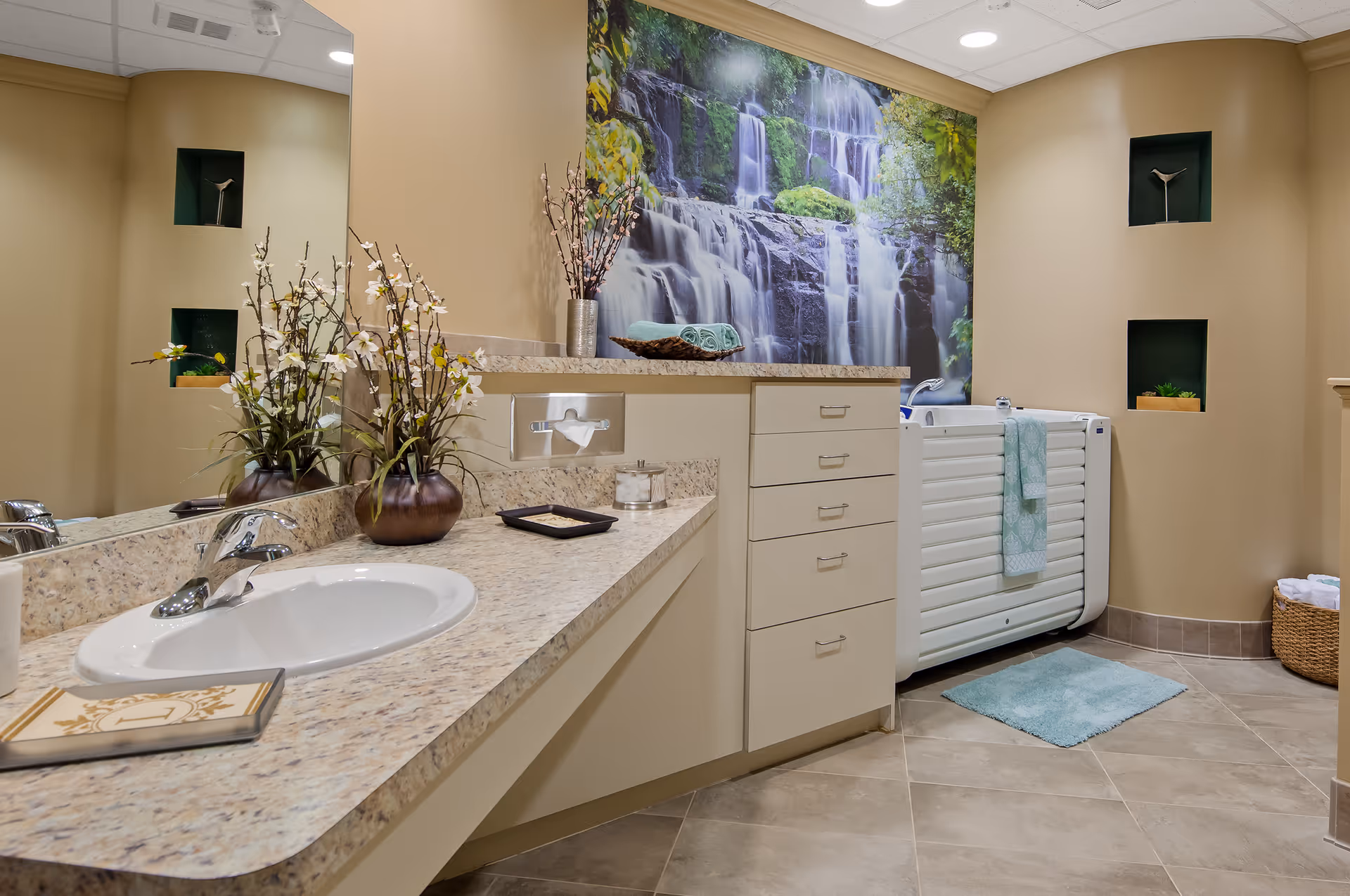 A clean and modern bathroom with a beige countertop and sink, decorated with a vase of white flowers. There is a large wall mural of a waterfall behind a white accessible bathtub with a teal towel hanging on it. The bathroom has beige walls with small recessed shelves holding decorative items and a basket with towels on the floor.
