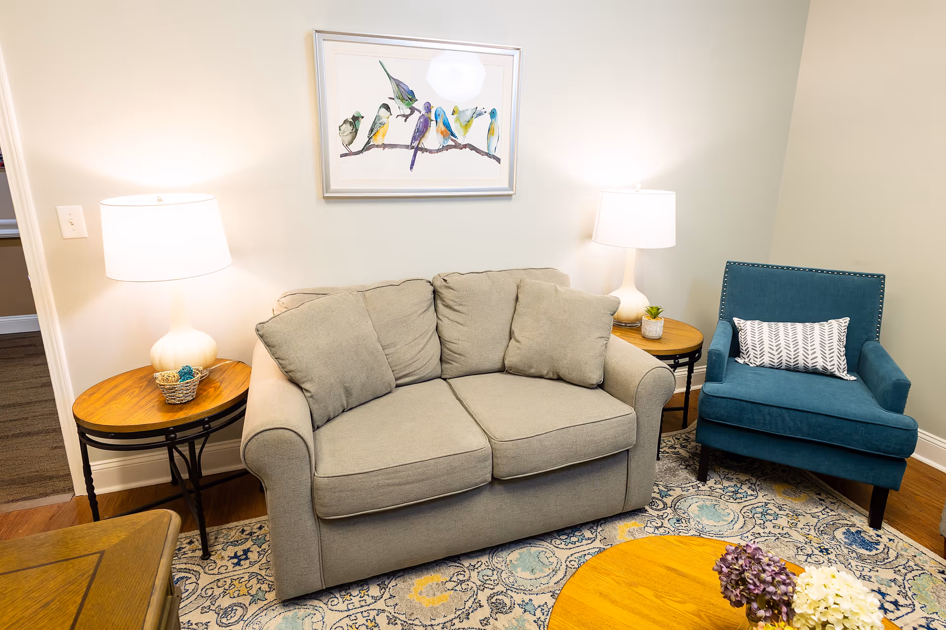 A cozy living room area with a light gray loveseat with three cushions, a blue armchair with a patterned pillow, two wooden side tables each with a white lamp, a framed picture of colorful birds on a branch hanging on the wall, and a round wooden coffee table with flower arrangements on a patterned rug.