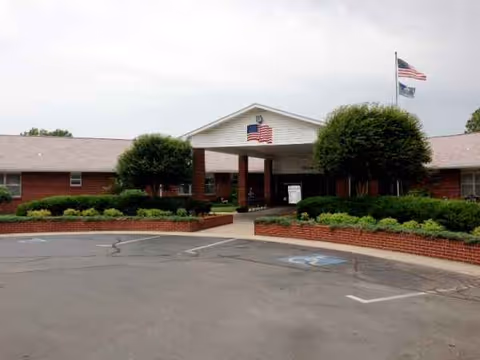Brick senior living facility front entrance with flagpole, manicured shrubs, and parking lot.
