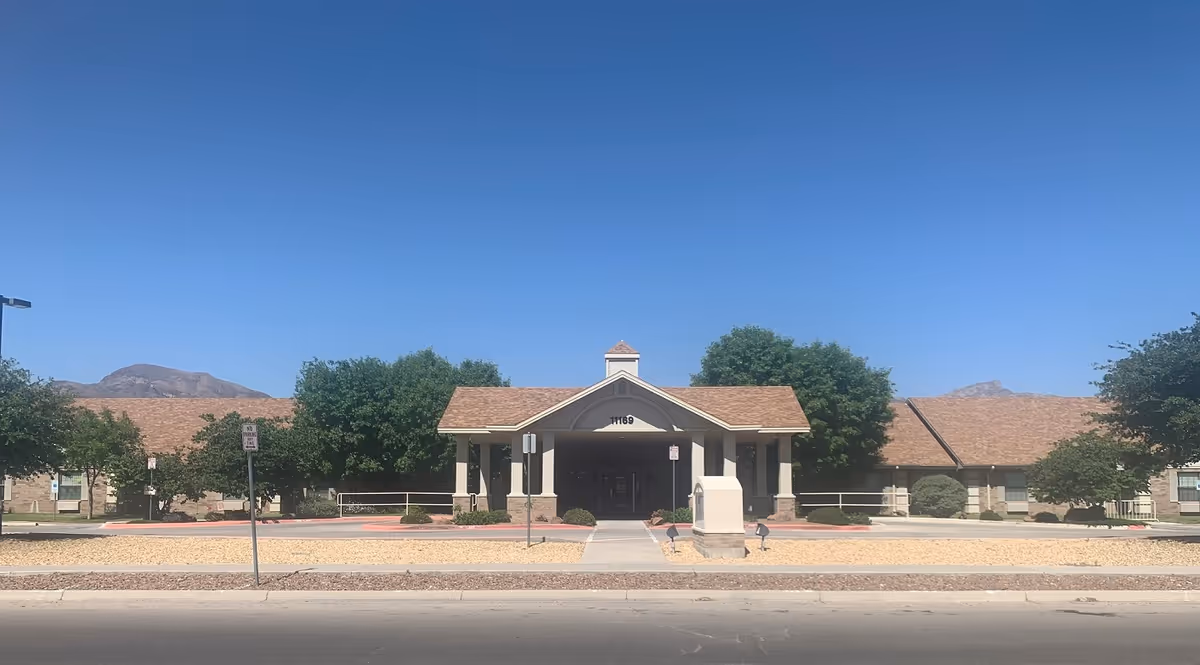 Front exterior view of Los Arcos del Norte Care Center building with a peaked roof entrance, surrounded by trees and landscaping under a clear blue sky.