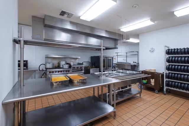 A clean commercial kitchen with stainless steel countertops and cooking equipment including ovens, stovetops, and warming trays. There are two trays of food on the counter, a rack filled with stacked plates, and a clock on the wall.