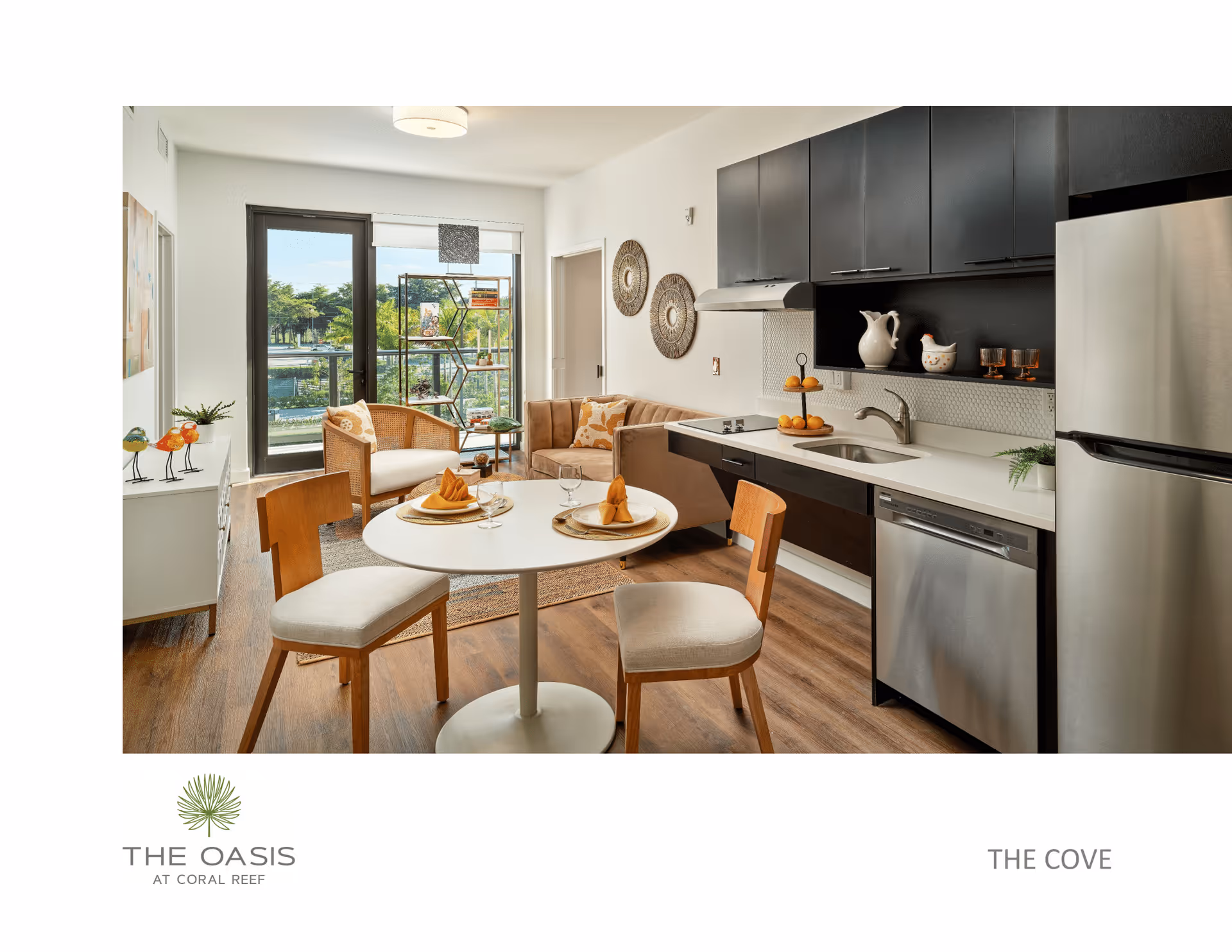 A modern open-plan living space featuring a small round dining table set for two with napkins and glasses, two wooden chairs with cushioned seats, a compact kitchen area with black cabinets, a stainless steel refrigerator, dishwasher, and sink. The living area includes a beige sofa with patterned cushions, a wicker armchair, a decorative shelving unit near a glass door leading to a balcony with a view of greenery and water outside. The room has wooden flooring and white walls with decorative wall hangings.