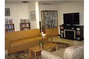 A cozy living room with a mustard yellow sofa, a patterned armchair, a wooden coffee table with a potted orchid, a bookshelf filled with books, and a flat-screen TV on a black stand with a fireplace below it.