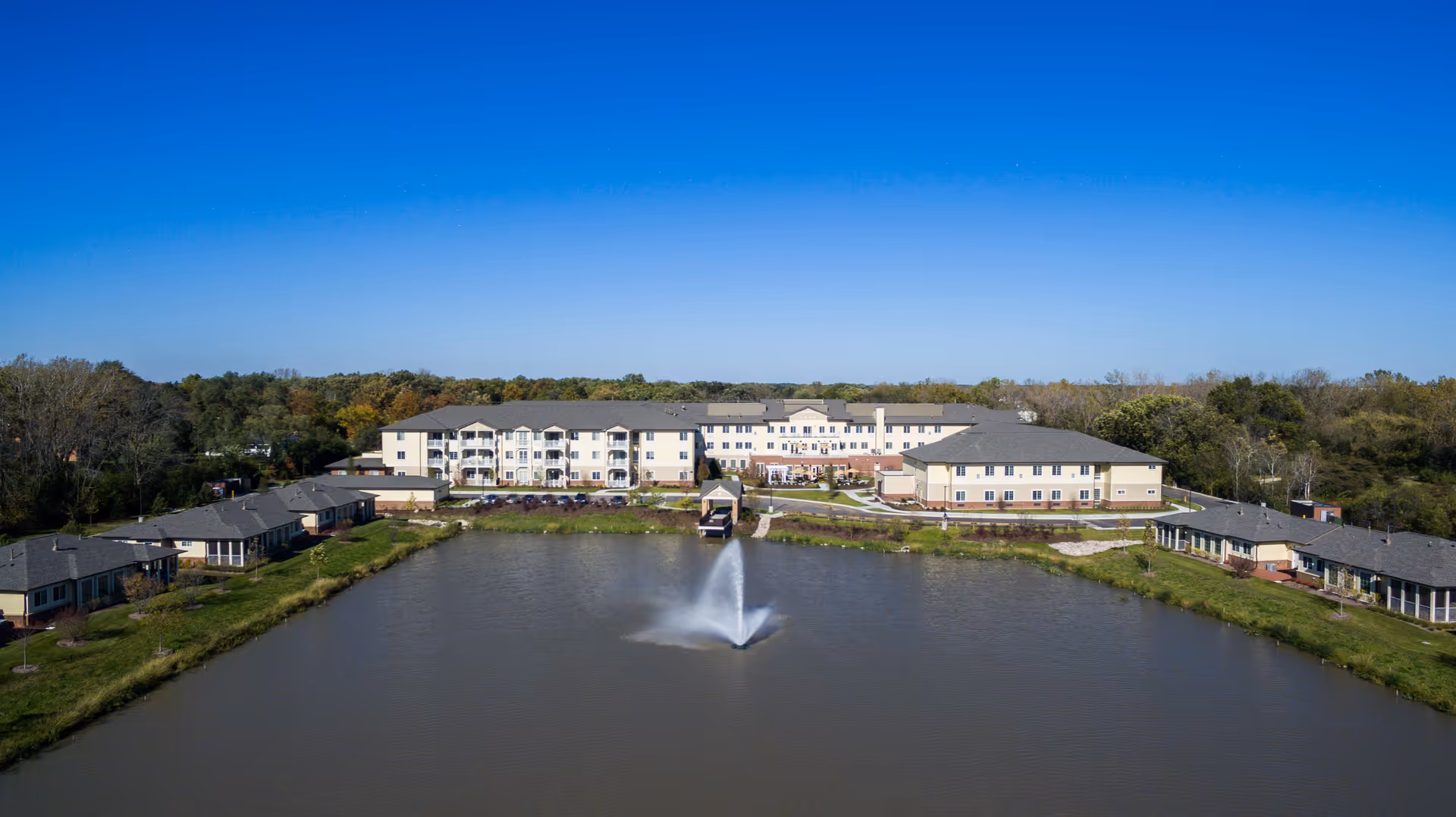Aerial view of Green Oaks Senior Living facility showing a large building complex with multiple wings surrounding a pond with a central water fountain, set against a backdrop of trees and clear blue sky.