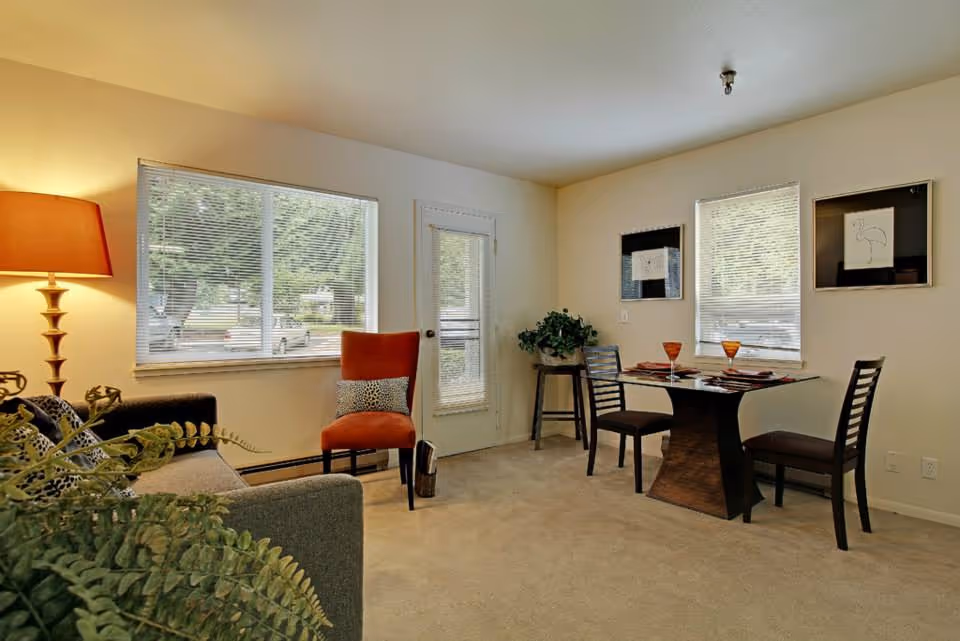 A cozy living and dining area with a gray sofa, an orange chair, a wooden dining table set for two with orange glasses, two windows with blinds, a door with a window, a floor lamp with an orange shade, and some green plants.