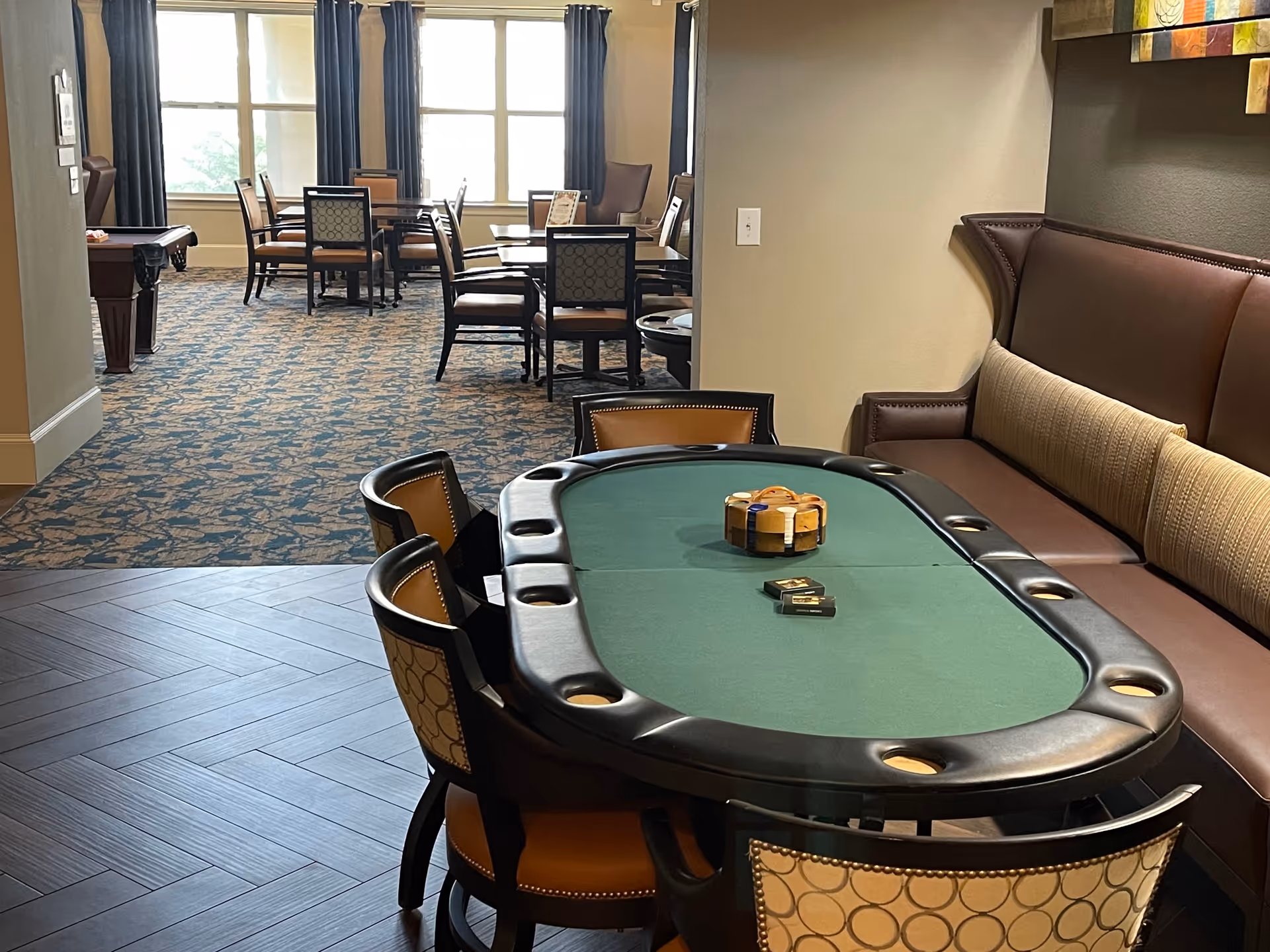 Interior view of a senior living facility game and dining area featuring a green felt poker table with chairs and a cushioned bench, poker chips and cards on the table, and several dining tables and chairs near large windows with curtains in the background.