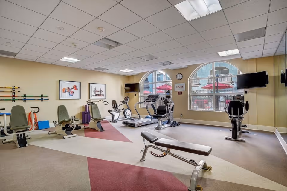 A fitness room with exercise equipment including stationary bikes, a treadmill, and a weight bench. The room has large arched windows showing an outdoor patio with red umbrellas. The walls are light yellow, and there are framed pictures and a clock on the wall. Two flat-screen TVs are mounted on the walls.
