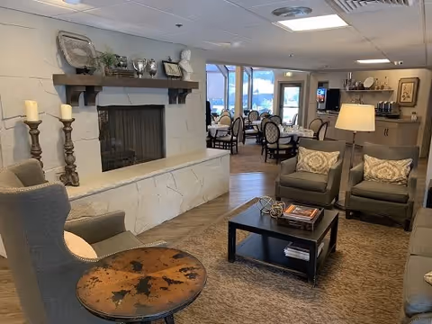 A cozy senior living facility common area featuring a stone fireplace with decorative items on the mantle, comfortable armchairs with patterned pillows, a coffee table with books and decor, and a dining area with tables and chairs visible in the background near large windows.