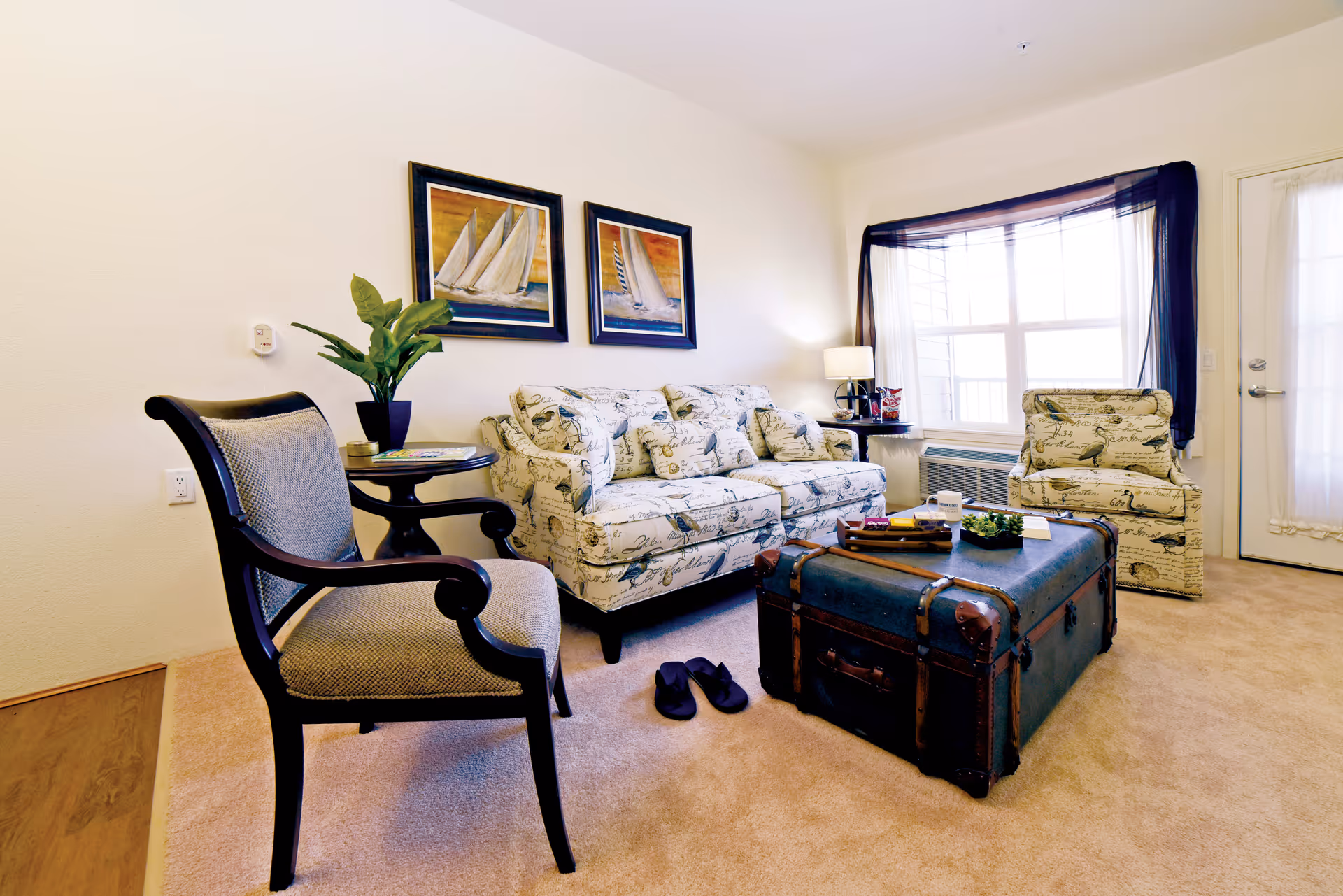 A cozy living room with a patterned sofa and matching armchair, a wooden framed chair with cushioned seat, a vintage trunk used as a coffee table, a small round side table with a plant, two framed sailboat paintings on the wall, a window with sheer black curtains, and a door with a sheer curtain.