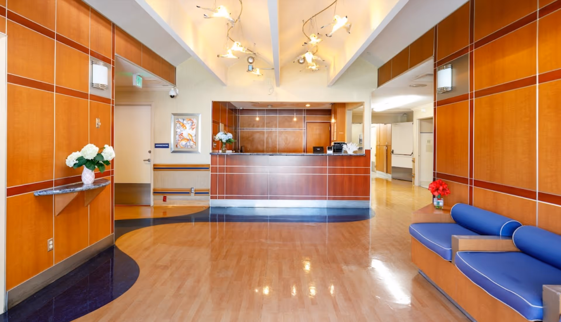 Reception area of Ocean Pointe Healthcare Center with a wooden front desk, blue cushioned seating on the right, wooden paneled walls, and decorative flowers on shelves. The floor is a combination of light wood and dark blue tiles, and the ceiling has modern light fixtures.