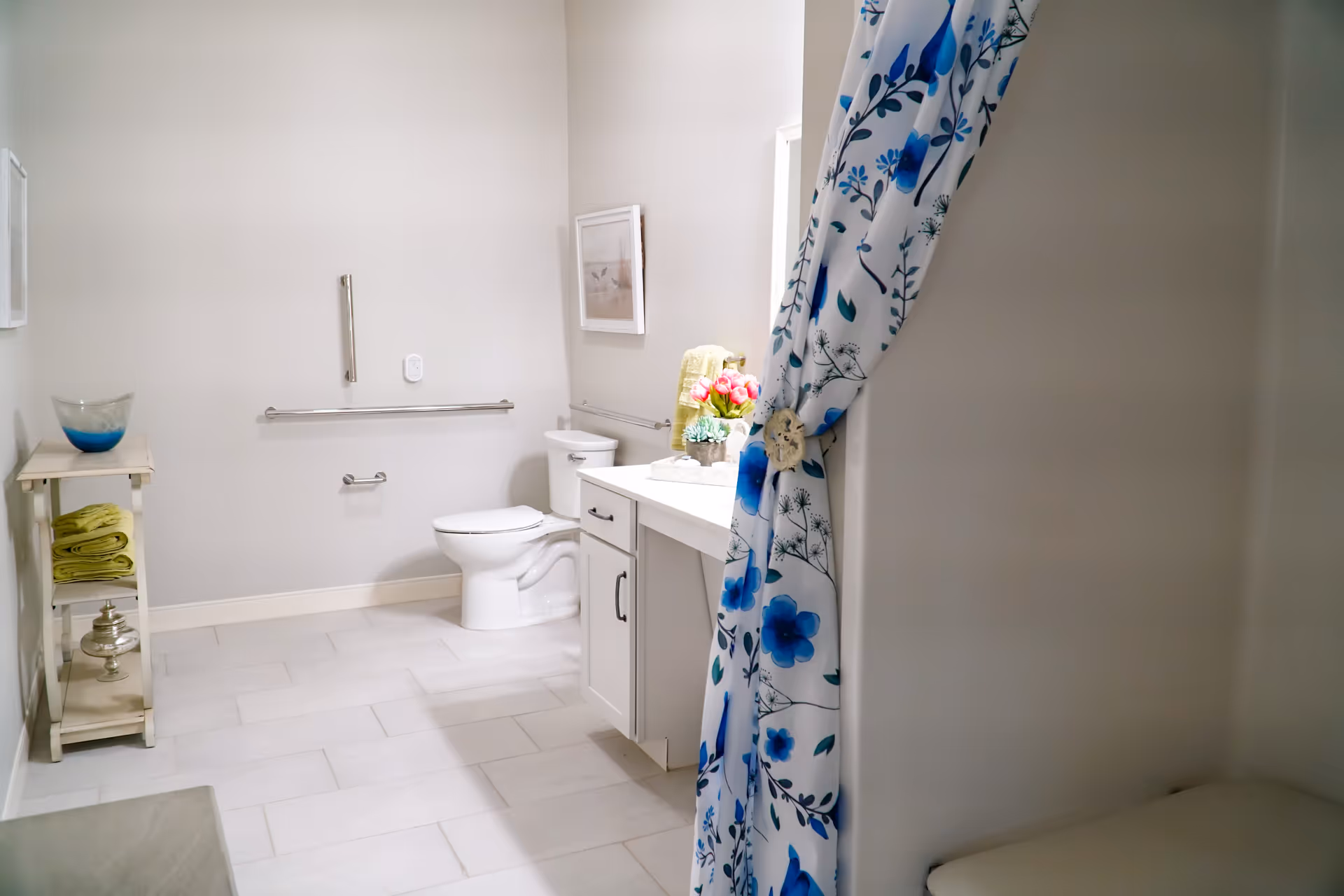 A clean and bright bathroom with a white toilet, a vanity with a sink, and a floral blue and white shower curtain tied to the side. There is a small shelf with green towels and decorative items, a framed picture on the wall, and grab bars near the toilet for accessibility.