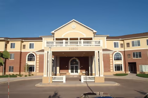 Front exterior view of a multi-story senior living facility building with a covered entrance and the number 14500 displayed above the entrance. The building features a combination of beige and brick facade with multiple windows and a clear blue sky in the background.