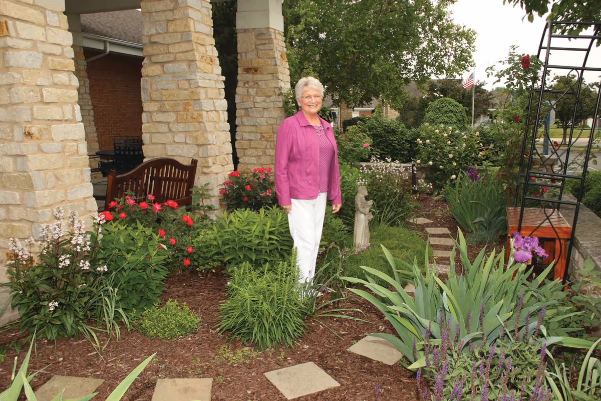An elderly woman wearing a purple jacket and white pants stands smiling in a well-maintained garden with various flowering plants and shrubs. Behind her are stone pillars supporting a covered porch with a wooden bench. An American flag is visible in the background.
