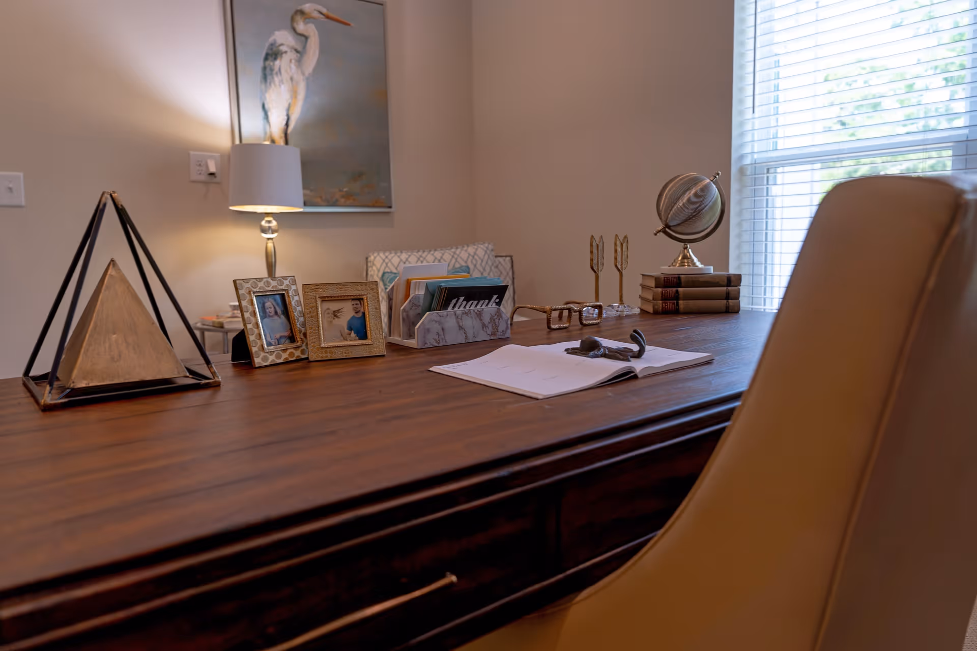 A wooden desk with decorative items including a triangular sculpture, two framed photos, a lamp, a card holder with cards, a pair of glasses, an open book with a small sculpture on it, two arrow-shaped bookends holding stacked books, and a globe near a window with blinds. A beige chair is partially visible in the foreground.