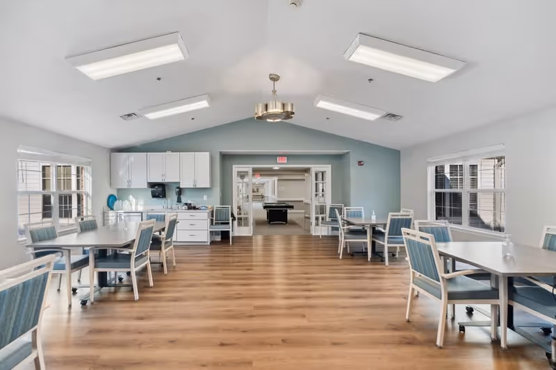 A bright and spacious dining area with multiple tables and chairs arranged neatly. The room has large windows on both sides allowing natural light to fill the space. At the far end, there is a kitchenette with white cabinets and a countertop. Beyond the dining area, through double glass doors, a room with a pool table is visible. The floor is wooden, and the ceiling has modern light fixtures.