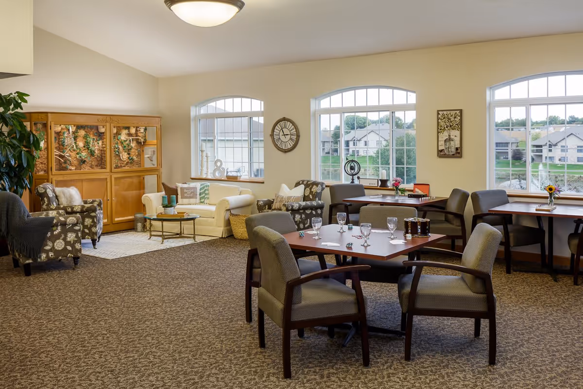 A spacious and well-lit common area in a senior living facility with multiple seating arrangements including armchairs, sofas, and tables with chairs. Large windows provide natural light and a view of residential buildings and greenery outside. The room features a carpeted floor, a wall clock, decorative items, and a large wooden enclosure with plants inside.