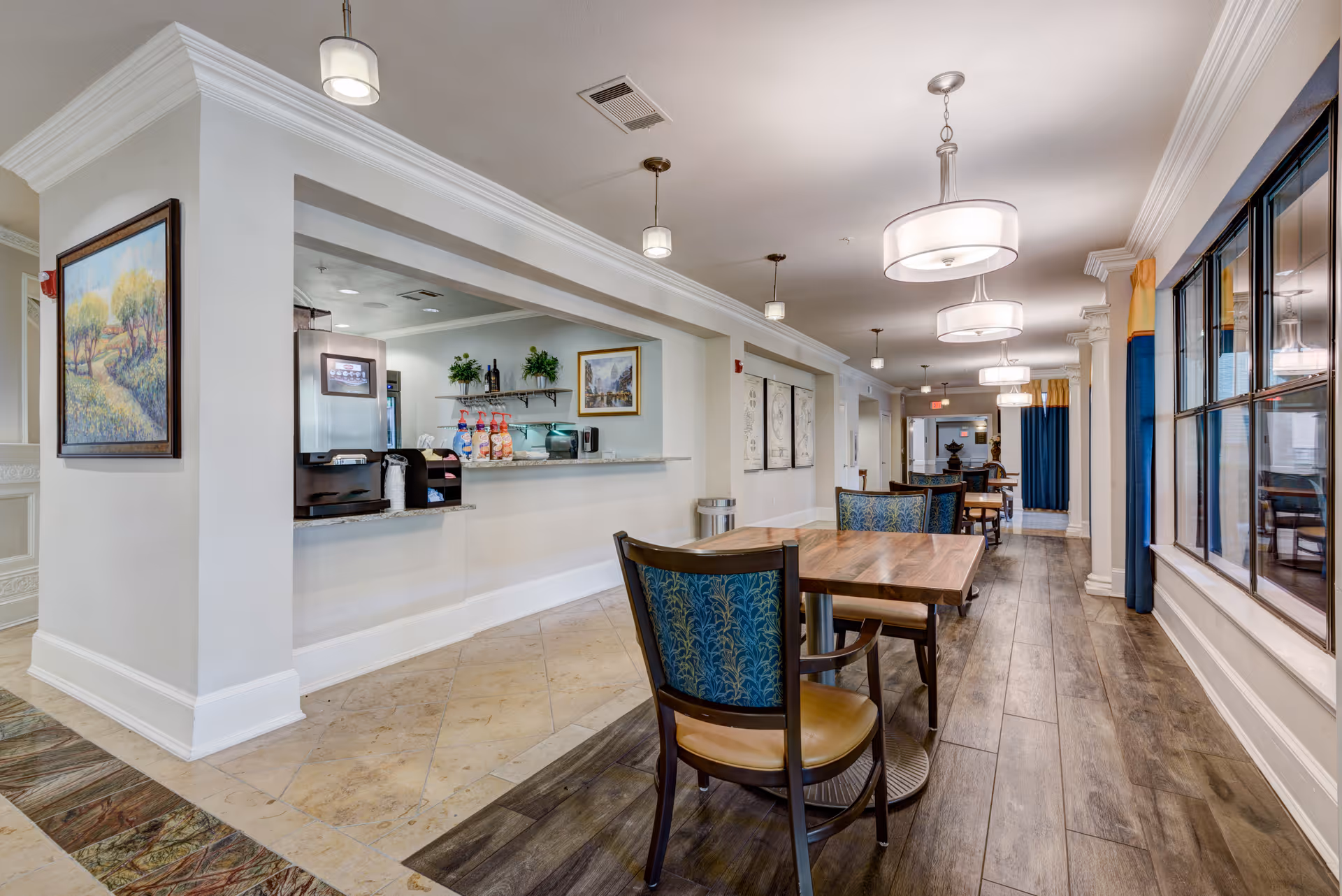 Interior view of a senior living facility dining area with wooden tables and chairs, pendant and ceiling lights, large windows with blue and yellow curtains, and a serving counter with coffee and condiments. The space has a mix of tile and wood flooring, white walls, and framed artwork.