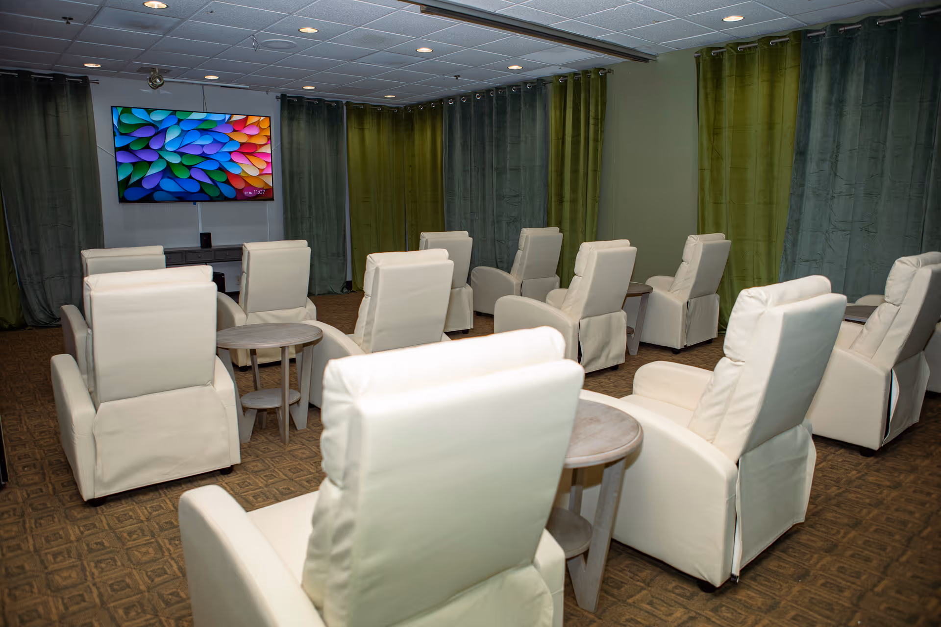 A room with multiple white recliner chairs arranged in rows facing a large wall-mounted flat screen TV displaying a colorful abstract image. Each pair of chairs is separated by small round wooden tables. The room has green curtains covering the walls and a patterned carpeted floor.