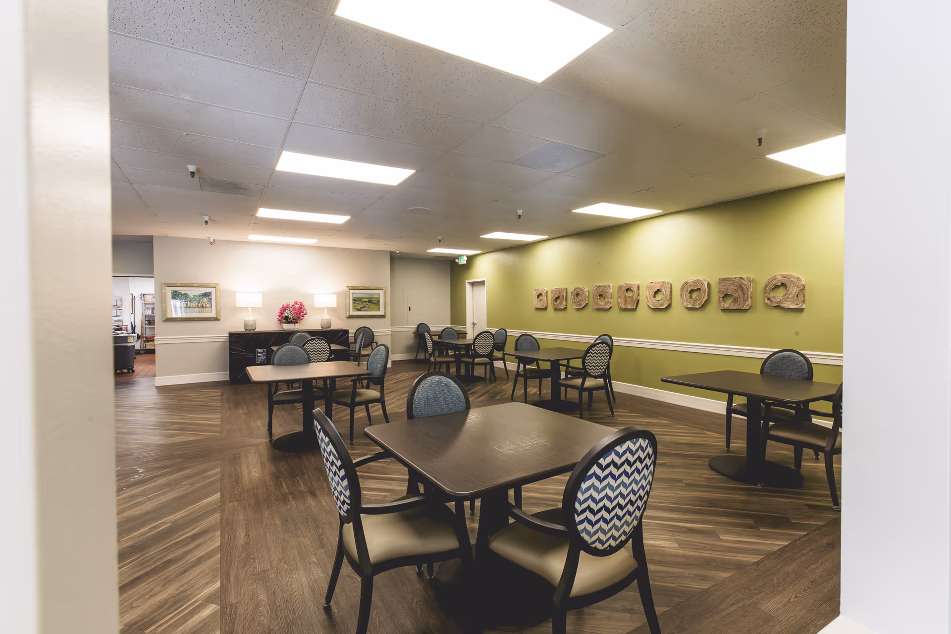 A dining room with several dark wood tables and chairs with patterned upholstery. The room has wood flooring, a green accent wall with decorative wooden art pieces, and two framed paintings on a white wall. The ceiling has recessed lighting, and there is a sideboard with two lamps and a flower arrangement.