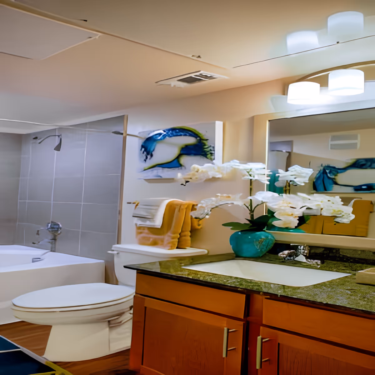 A bathroom with a white toilet, a bathtub with a showerhead, a wooden vanity with a granite countertop, a sink, a large mirror, and a turquoise vase with white flowers. There are folded towels on a rack above the toilet and a colorful abstract painting on the wall.