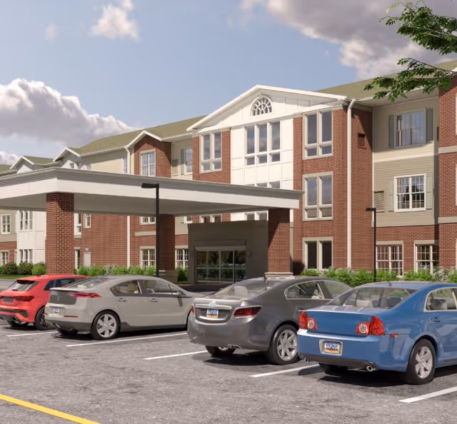Exterior view of a senior living facility named Harmony at Glasgow, showing a multi-story brick and beige building with large windows, a covered entrance, and a parking lot with several parked cars under a partly cloudy sky.