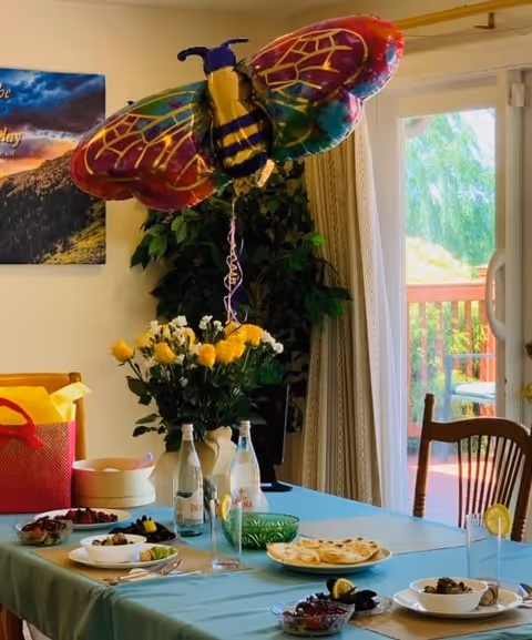 A dining table set with plates of food, glasses with lemon slices, and bottles of water. A large colorful butterfly balloon is tied to a vase filled with yellow and white flowers. In the background, there is a sliding glass door leading to a wooden deck with outdoor greenery visible. A framed picture hangs on the wall to the left.