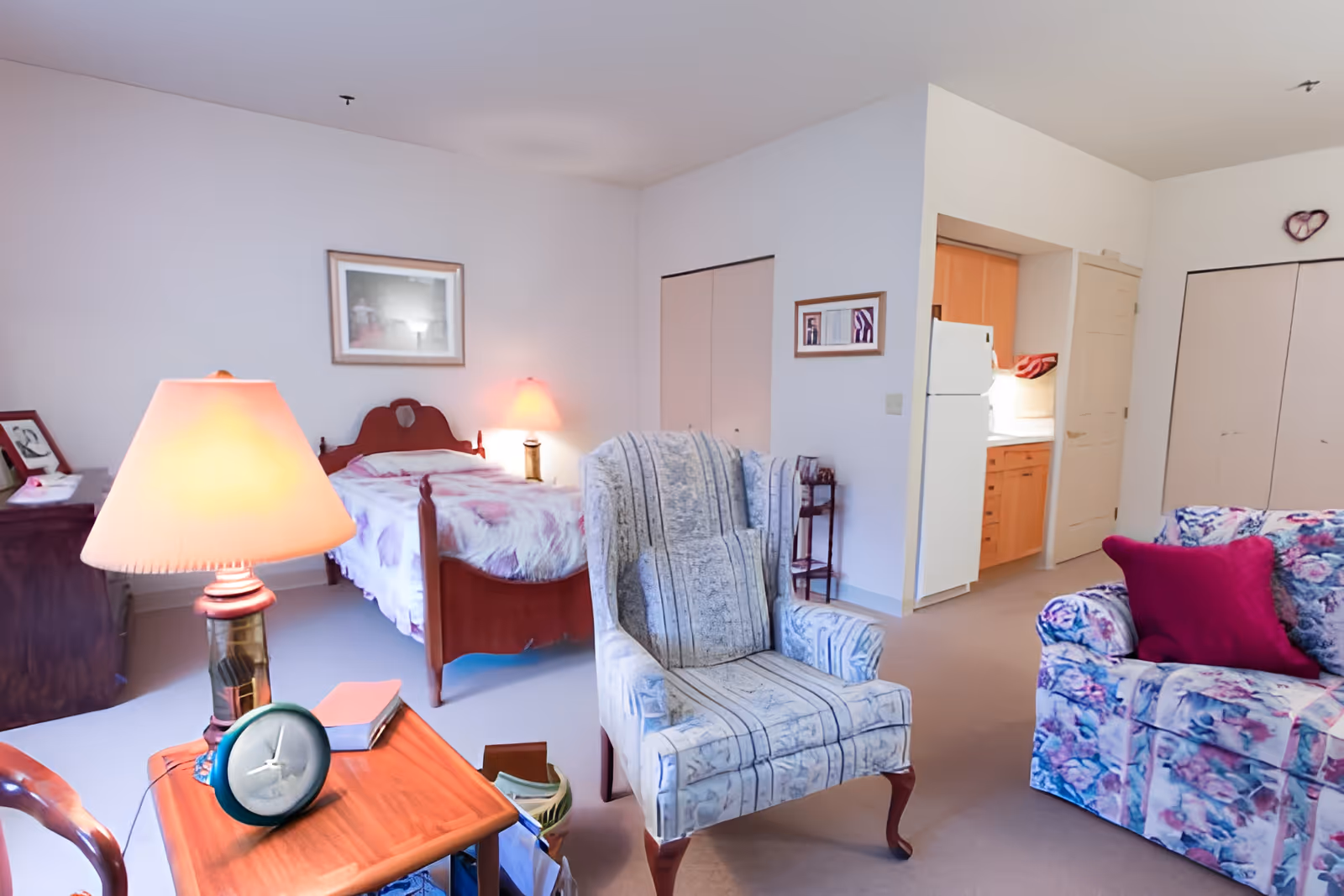 A cozy senior living room and bedroom area with a floral-patterned armchair and sofa, a wooden side table with a lamp and clock, a wooden bed with a floral bedspread, and a small kitchen area visible in the background.
