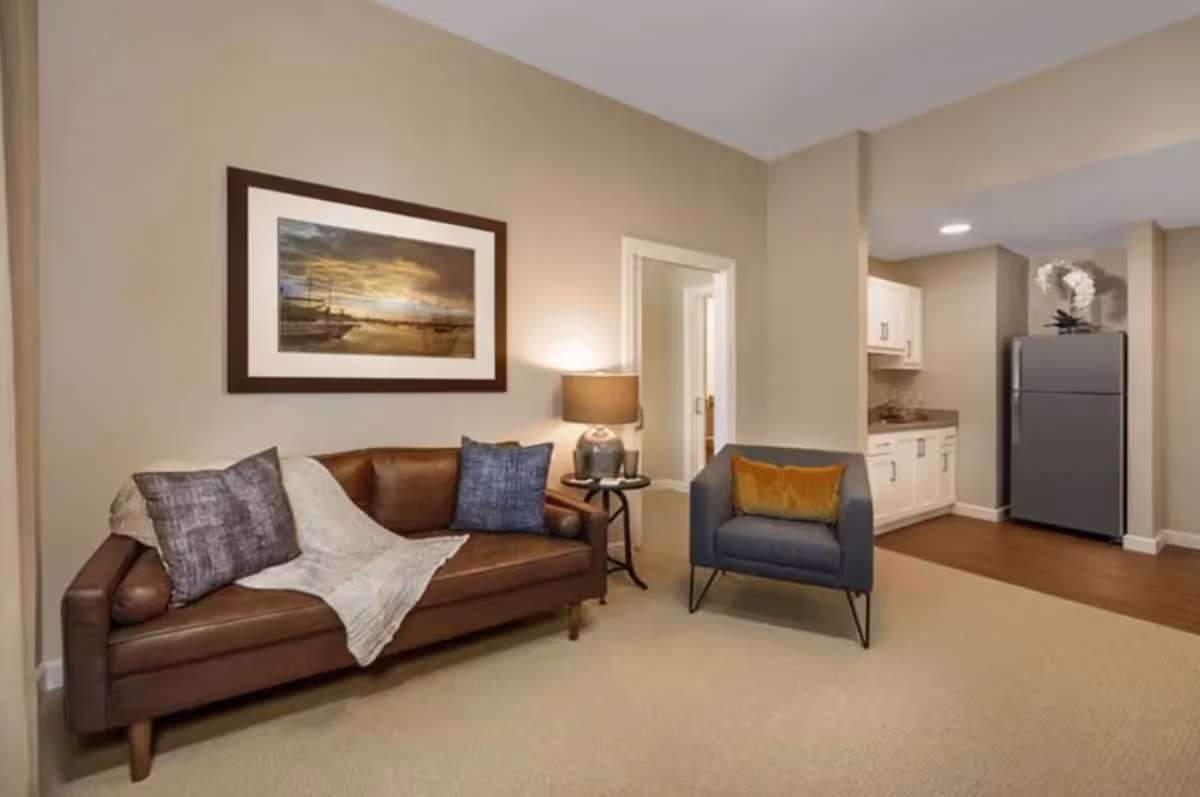 A cozy living room area featuring a brown leather sofa with two pillows and a light throw blanket, a small round side table with a lamp, and a gray armchair with an orange cushion. In the background, there is a kitchenette with white cabinets and a stainless steel refrigerator. The walls are painted beige, and a framed picture of a sunset over boats hangs above the sofa.