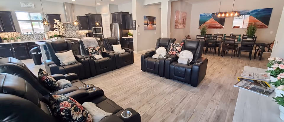 Spacious interior room featuring a row of black leather recliner chairs with floral and light-colored pillows arranged facing a kitchen area. The kitchen has dark cabinets, a mosaic tile backsplash, and stainless steel appliances. In the background, there is a dining area with a long table, chairs, and decorative wall art. The floor is light wood, and the room is well-lit with natural and artificial lighting.