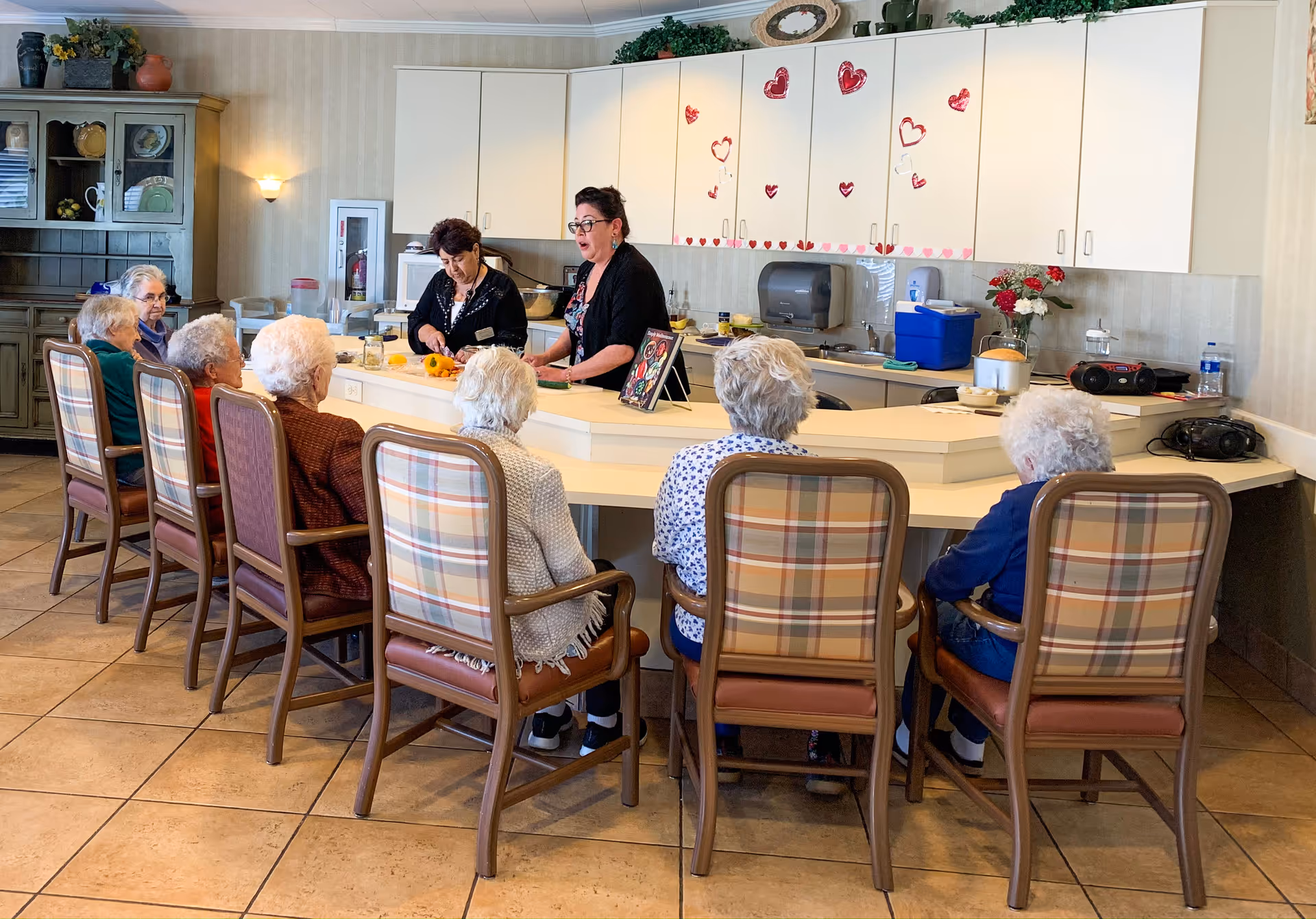 Several elderly residents sit around a curved counter while two staff members prepare food and lead an activity in a communal kitchen/dining area decorated with heart stickers.