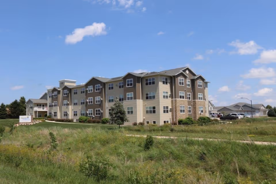 Three-story beige and brown senior living building with many windows and landscaped grounds under a blue sky.