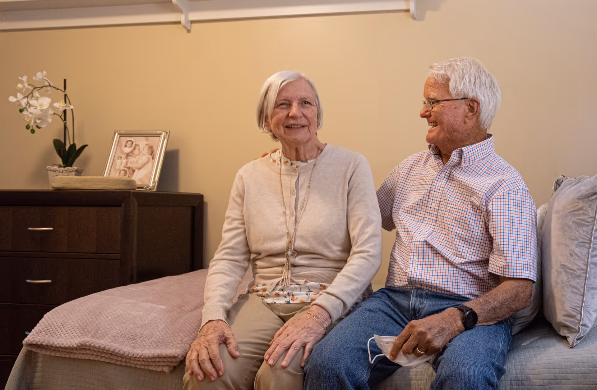 An elderly couple sitting on a bed in a cozy bedroom. The woman is wearing a beige cardigan and light-colored pants, while the man is wearing a checkered shirt and jeans, holding a face mask in his hand. Behind them is a wooden dresser with a framed photo and a white orchid plant.