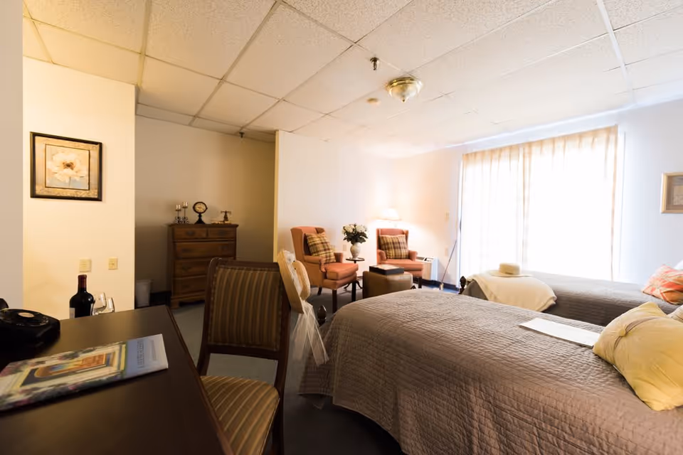 A cozy bedroom in a senior living facility with two twin beds covered in gray quilts, each with pillows. There are two upholstered armchairs with plaid cushions near a window with sheer curtains, a small table with a lamp and flowers between them. A wooden dresser with decorative items is visible in the background, along with a framed flower picture on the wall. A desk with a chair, a bottle of wine, and a magazine is in the foreground.