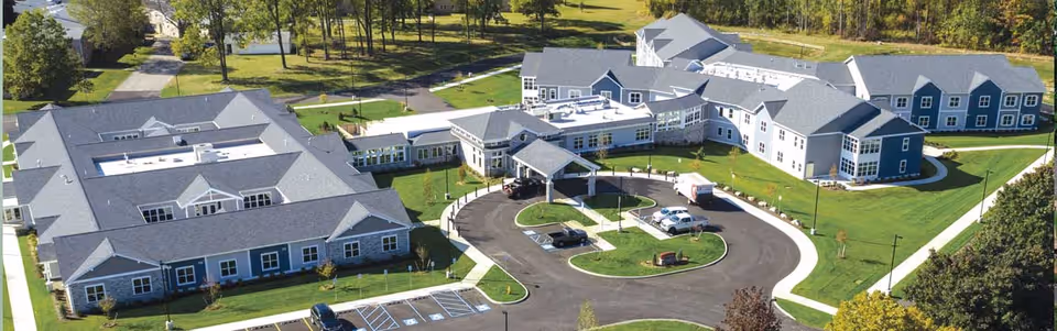Aerial view of the Brothers of Mercy senior living facility showing multiple connected buildings with gray roofs, surrounded by green lawns and trees. There is a circular driveway with parked cars and a covered entrance in the center of the complex.