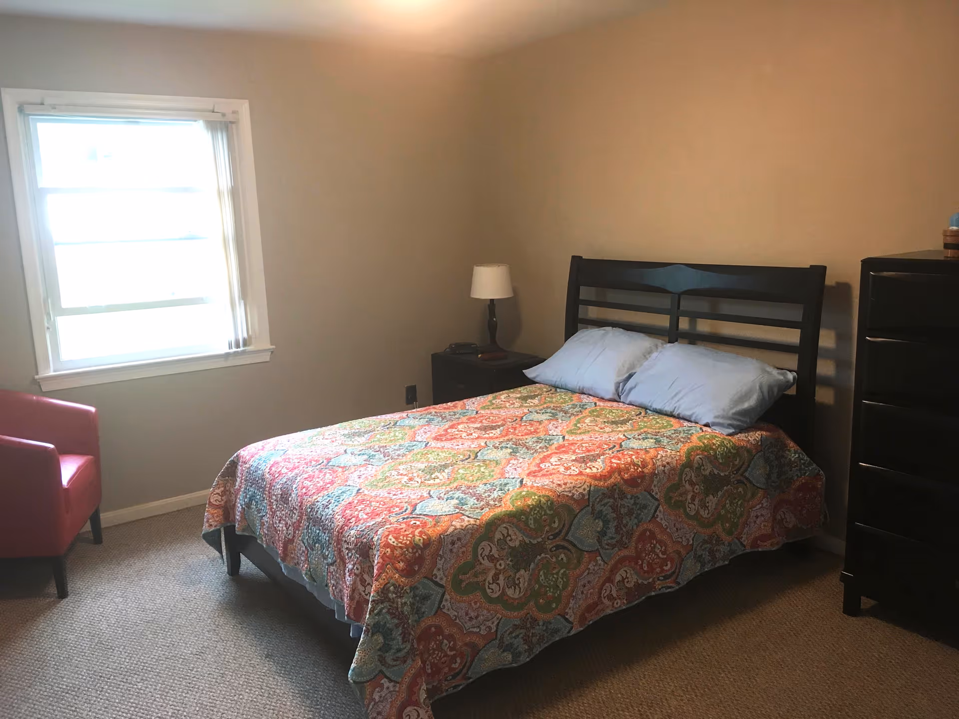A bedroom with a bed covered in a colorful patterned quilt and two light blue pillows. There is a black headboard, a black nightstand with a lamp and a phone, a black dresser, a red armchair, and a window with blinds letting in natural light.