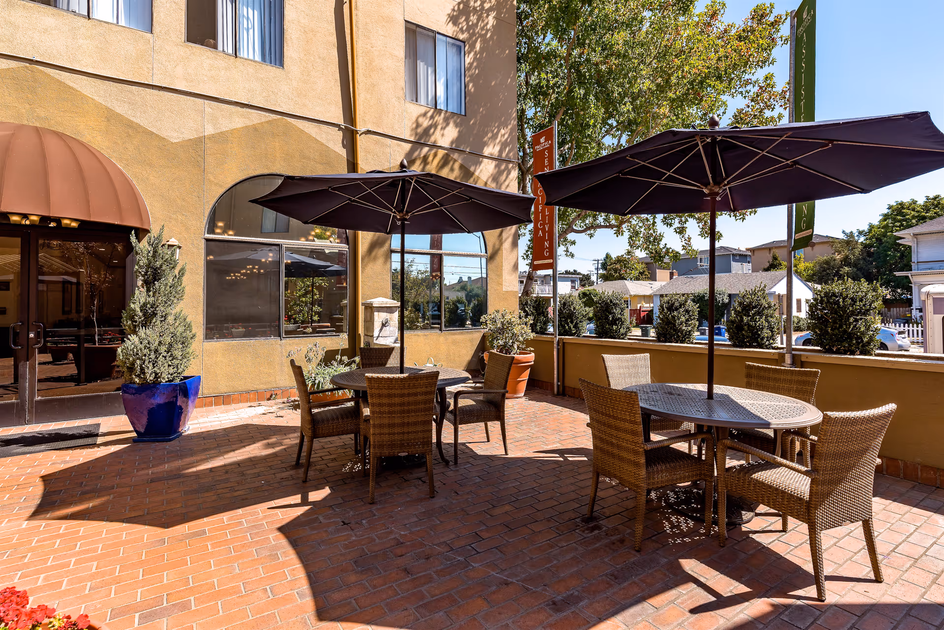 Outdoor brick patio with round tables, wicker chairs and large umbrellas outside a senior living building.