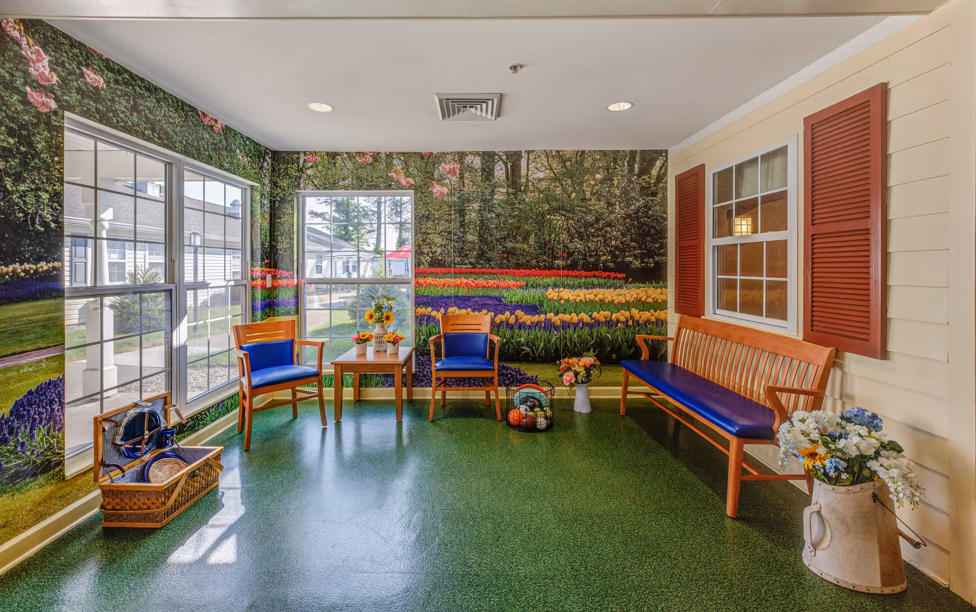 A bright indoor seating area with a green floor and walls decorated with large windows and a mural of a colorful flower garden. The room contains wooden chairs with blue cushions, a wooden bench with a blue cushion, a small wooden table with flower arrangements, a basket with a hat and other items, and a large vase with flowers. The wall on the right side has a window with red shutters.