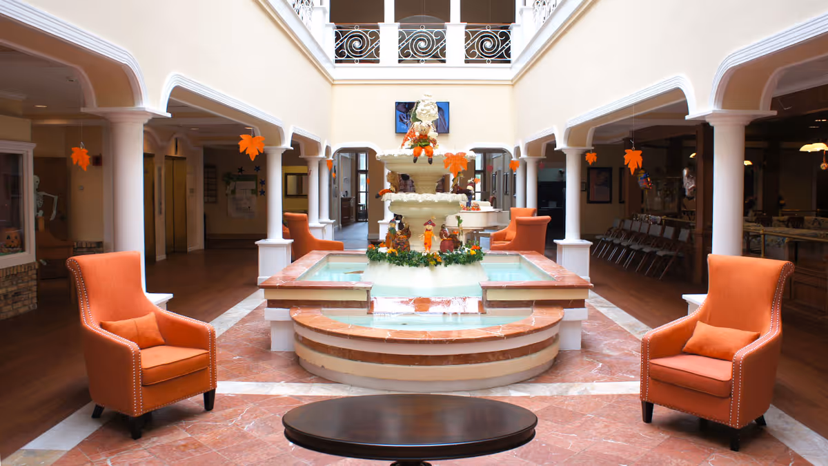 Interior view of a senior living facility lobby area with a central water fountain decorated with autumn-themed ornaments. The space features four orange armchairs arranged around the fountain, white columns, and a second-floor balcony with decorative railings. Hanging orange leaf decorations are visible from the ceiling.