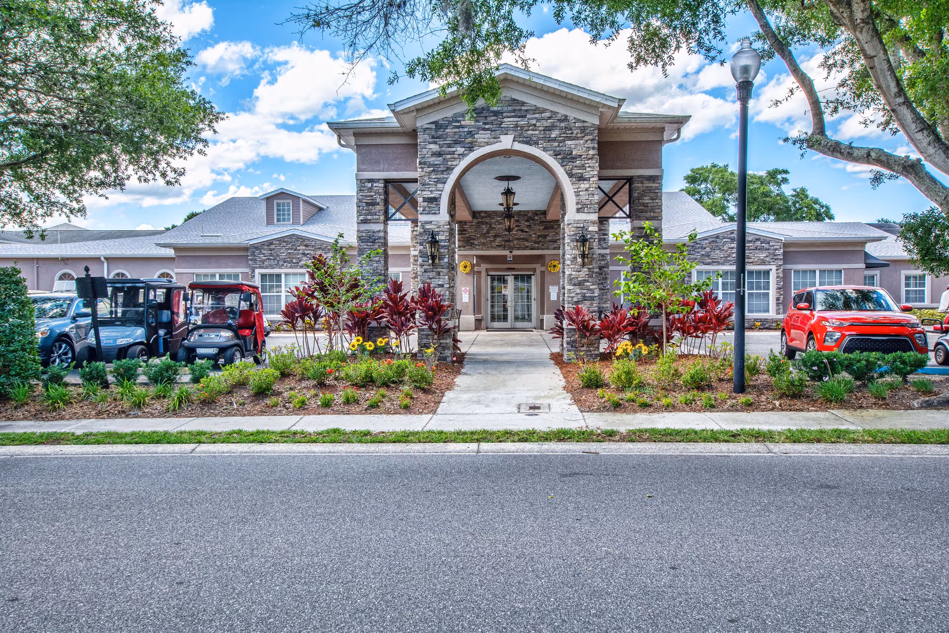 Stone-faced entrance of a retirement community building with an arched portico, landscaped flower beds and parked vehicles along the driveway.