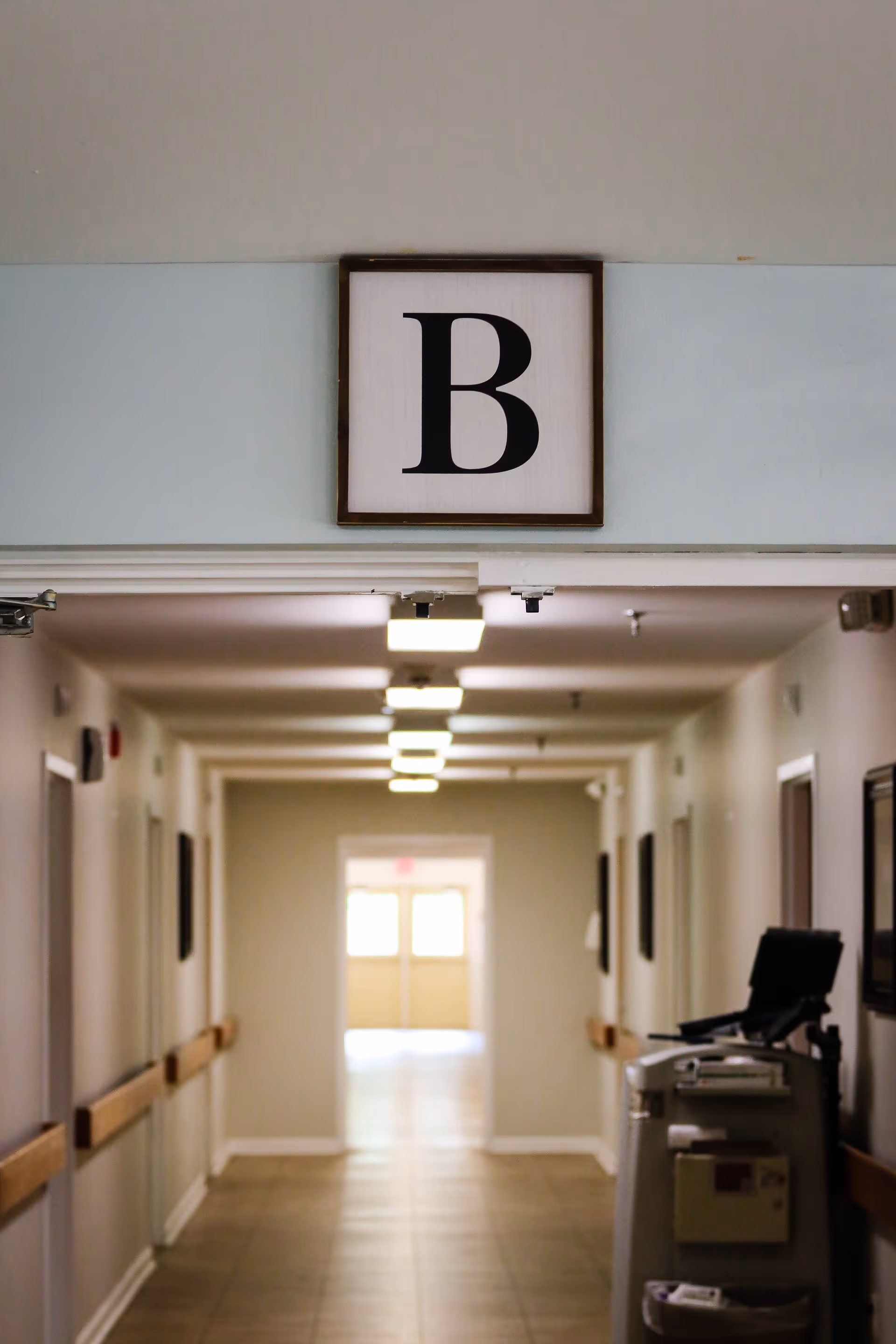 A well-lit hallway in a senior living facility with beige walls, handrails on both sides, and a sign with the letter B hanging above the entrance to the corridor. There is a medical cart or equipment on the right side of the hallway and double doors at the far end letting in natural light.