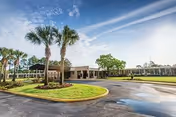 Exterior view of a single-story health and rehabilitation center building with a circular driveway, palm trees, and a well-maintained lawn under a partly cloudy sky.